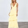 A woman modeling a pale yellow sleeveless maxi dress with a deep V-neckline, fitted bodice, and tiered ruffle skirt, standing against a light gray studio background.