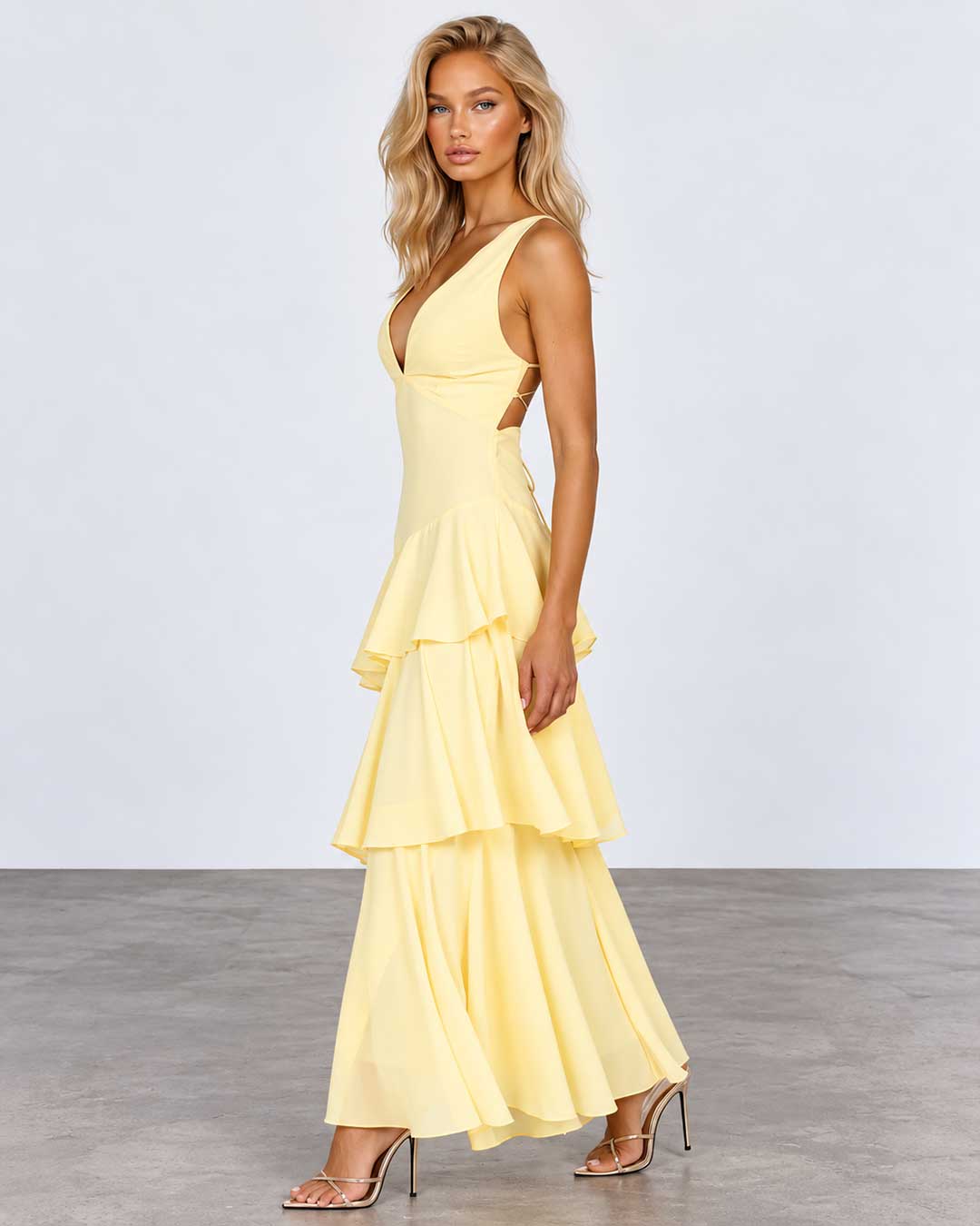 A woman modeling a pale yellow sleeveless maxi dress with a deep V-neckline, side cutouts, and a layered ruffle skirt, standing against a light gray studio background.