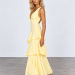 A woman modeling a pale yellow sleeveless maxi dress with a deep V-neckline, side cutouts, and a layered ruffle skirt, standing against a light gray studio background.