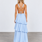 A woman stands with her back to the camera wearing a light blue tiered maxi dress with a strappy open back, paired with beige high heels, set against a plain studio background.