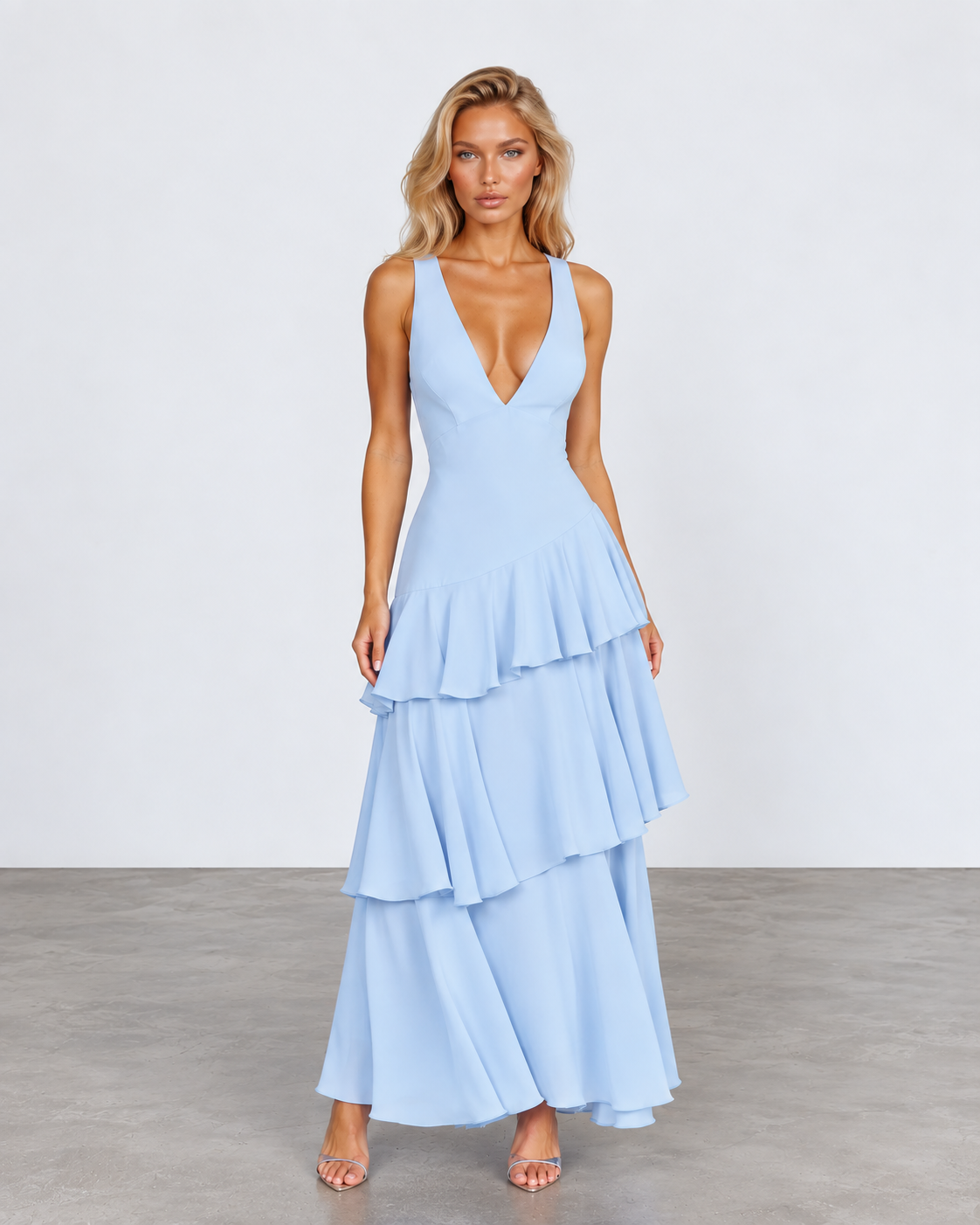 A woman modeling a light blue sleeveless maxi dress with a deep V-neckline and a tiered ruffle skirt, standing against a neutral studio backdrop.
