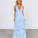 A woman modeling a light blue sleeveless maxi dress with a deep V-neckline and a tiered ruffle skirt, standing against a neutral studio backdrop.