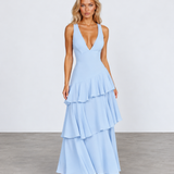 A woman modeling a light blue sleeveless maxi dress with a deep V-neckline and a tiered ruffle skirt, standing against a neutral studio backdrop.