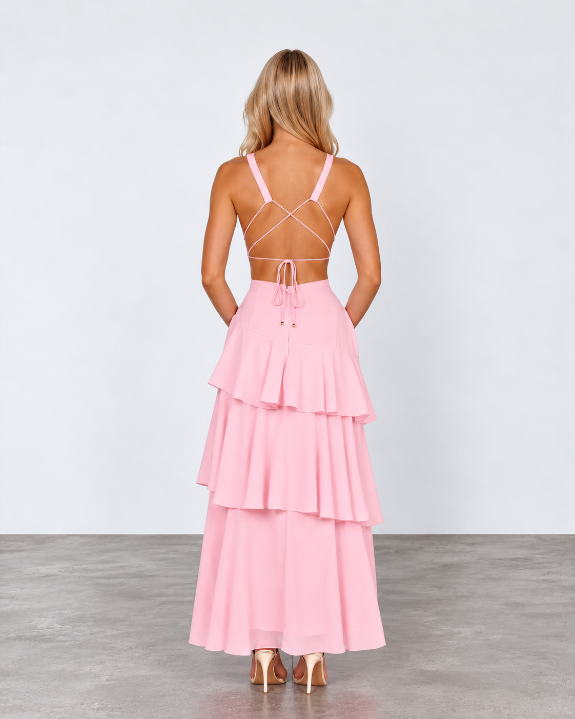 A woman stands with her back to the camera wearing a light pink tiered maxi dress with thin crisscross back straps and metallic gold high heels, set against a plain light gray studio background.