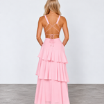 A woman stands with her back to the camera wearing a light pink tiered maxi dress with thin crisscross back straps and metallic gold high heels, set against a plain light gray studio background.
