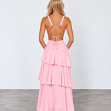 A woman stands with her back to the camera wearing a light pink tiered maxi dress with thin crisscross back straps and metallic gold high heels, set against a plain light gray studio background.
