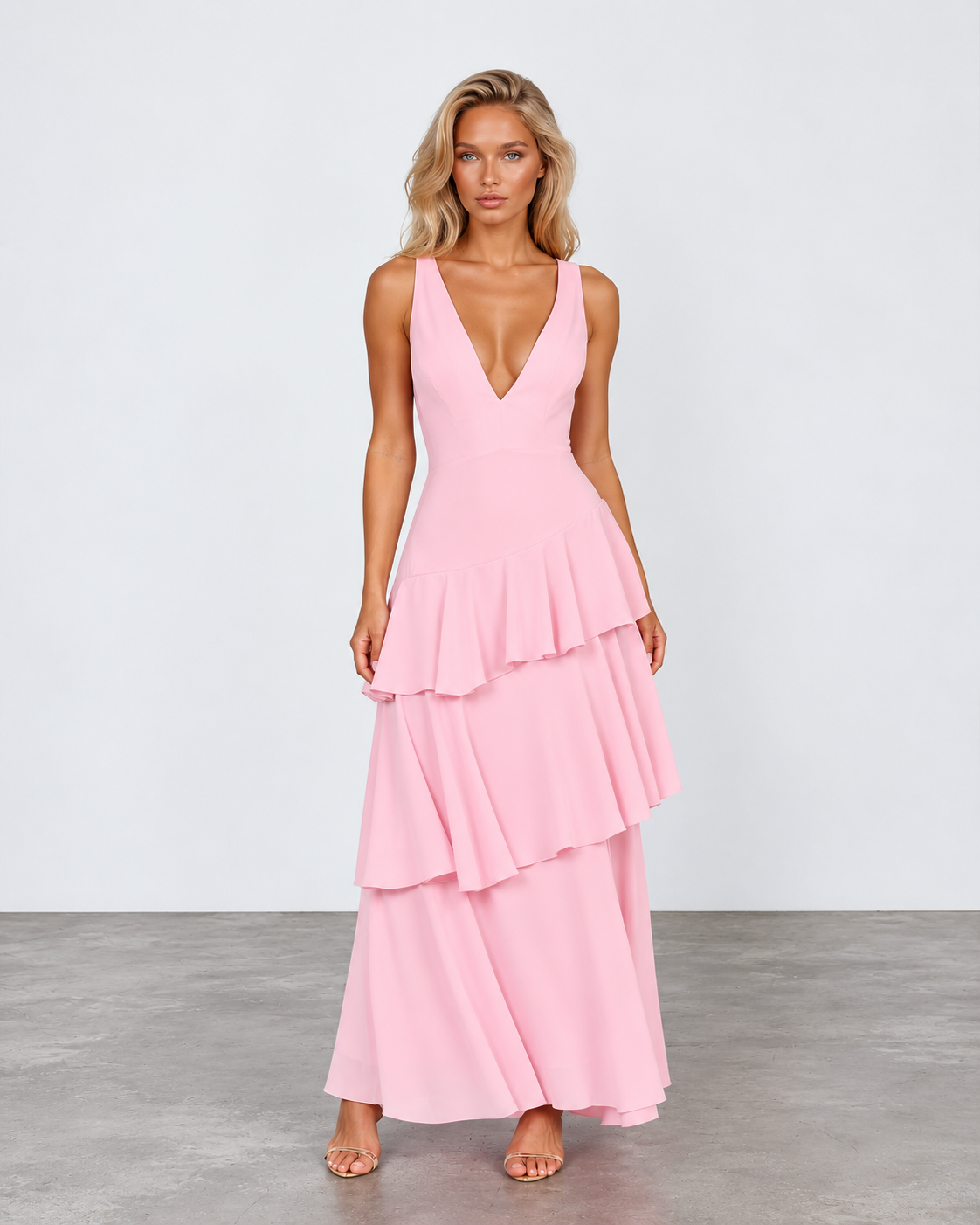 A woman modeling a sleeveless pink maxi dress with a deep V-neckline and a tiered ruffle skirt, standing against a light gray studio background.