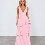 A woman modeling a sleeveless pink maxi dress with a deep V-neckline and a tiered ruffle skirt, standing against a light gray studio background.