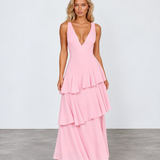 A woman modeling a sleeveless pink maxi dress with a deep V-neckline and a tiered ruffle skirt, standing against a light gray studio background.