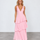 A woman modeling a sleeveless pink maxi dress with a deep V-neckline and a tiered ruffle skirt, standing against a light gray studio background.