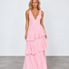 A woman modeling a sleeveless pink maxi dress with a deep V-neckline and a tiered ruffle skirt, standing against a light gray studio background.