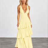 A woman modeling a pale yellow sleeveless maxi dress with a deep V-neckline and a tiered ruffle skirt, standing against a light gray studio background.