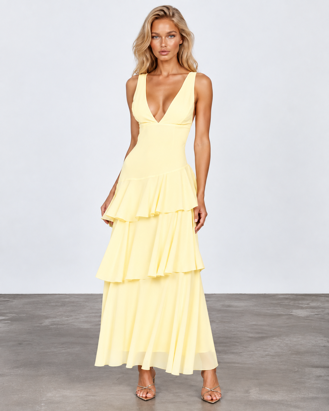A woman modeling a pale yellow sleeveless maxi dress with a deep V-neckline and a tiered ruffle skirt, standing against a light gray studio backdrop.