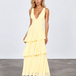 A woman modeling a pale yellow sleeveless maxi dress with a deep V-neckline and a tiered ruffle skirt, standing against a light gray studio backdrop.