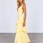 A woman modeling a pale yellow sleeveless maxi dress with a deep V-neckline, side cutouts, and a layered ruffle skirt, standing against a light gray studio background.