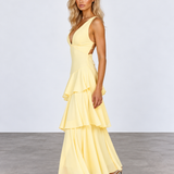 A woman modeling a pale yellow sleeveless maxi dress with a deep V-neckline, side cutouts, and a layered ruffle skirt, standing against a light gray studio background.