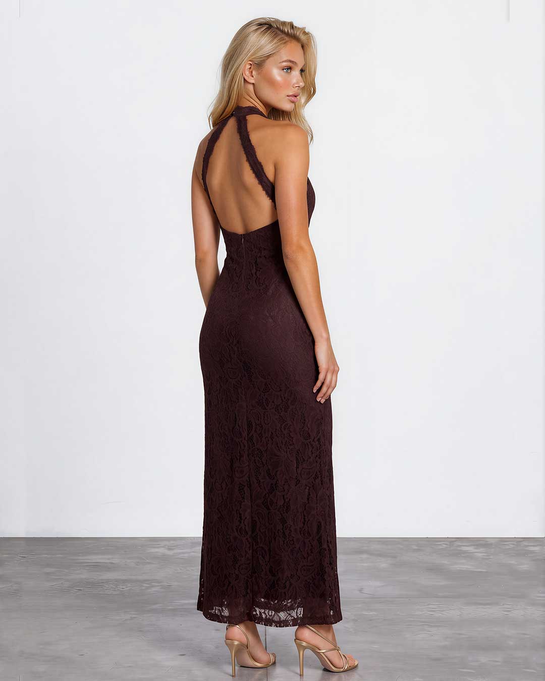A woman modeling a dark burgundy lace maxi dress with a halter-style open back, shown from the back in a studio setting with a white wall and gray floor.