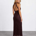 A woman modeling a dark burgundy lace maxi dress with a halter-style open back, shown from the back in a studio setting with a white wall and gray floor.