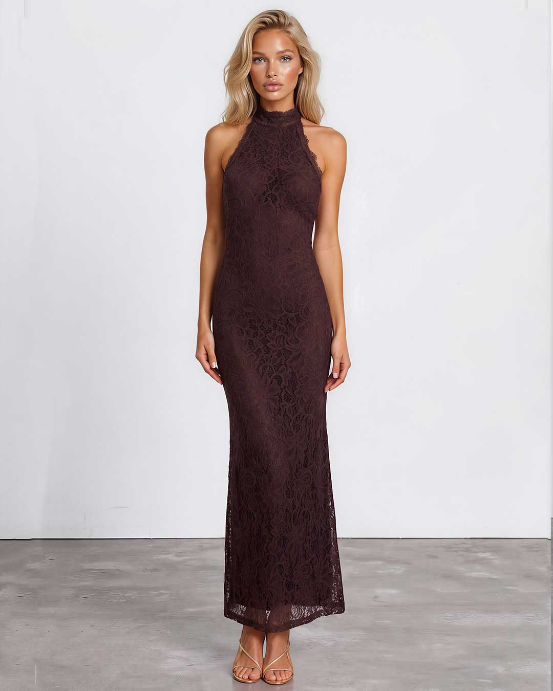 A woman modeling a sleeveless, high-neck, floor-length lace dress in a deep dark brown color, standing against a white studio wall.