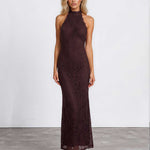 A woman modeling a sleeveless, high-neck, floor-length lace dress in a deep dark brown color, standing against a white studio wall.