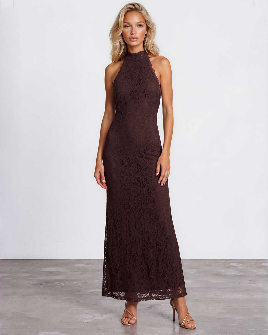 A woman modeling a sleeveless, high-neck, floor-length lace maxi dress in a deep dark brown color, standing against a light gray studio background.