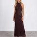 A woman modeling a sleeveless, high-neck, floor-length lace maxi dress in a deep dark brown color, standing against a light gray studio background.