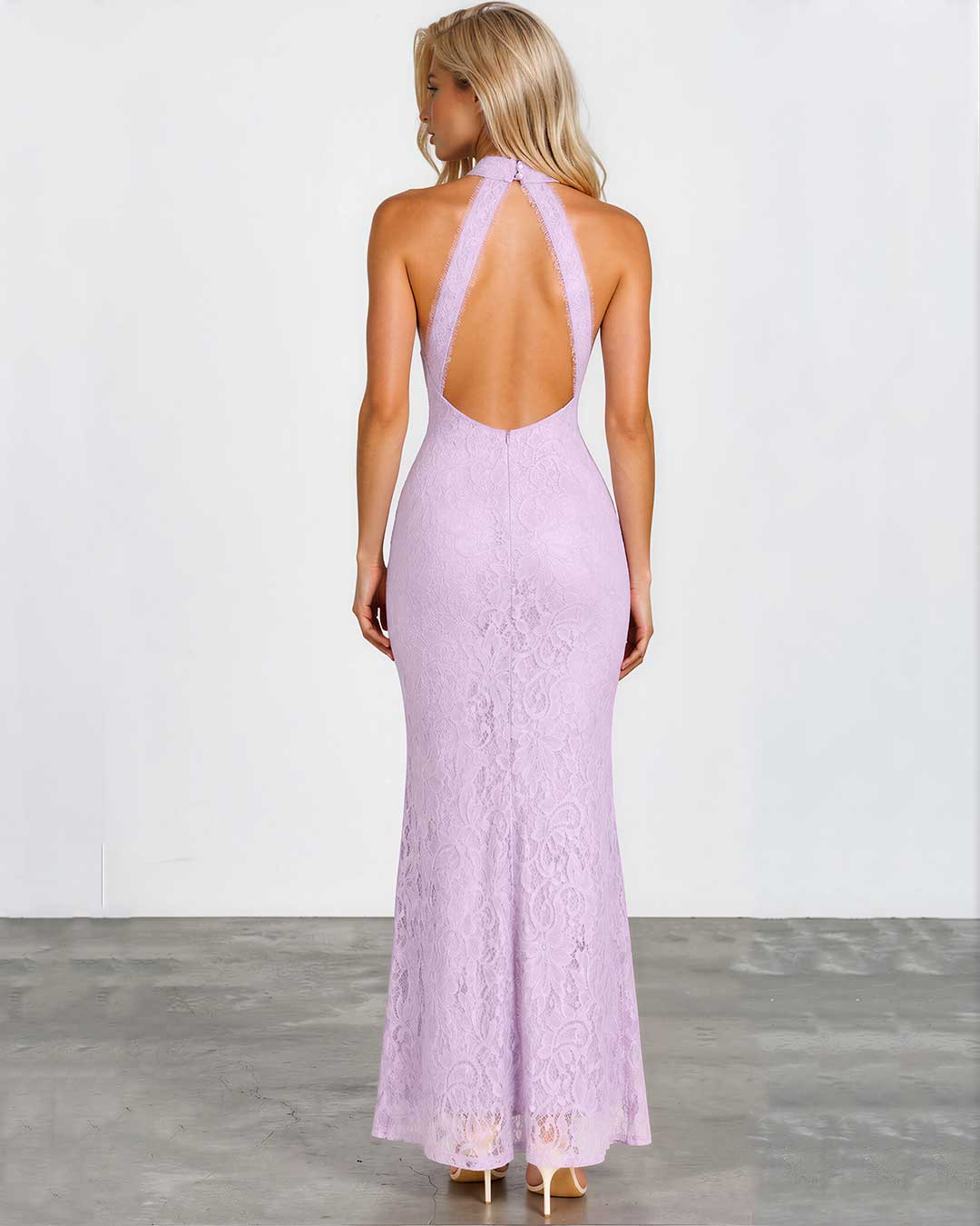 A woman viewed from the back wearing a lavender lace halter maxi dress with a deep open back, standing against a plain light gray wall and concrete floor.