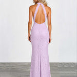 A woman viewed from the back wearing a lavender lace halter maxi dress with a deep open back, standing against a plain light gray wall and concrete floor.