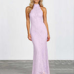 A woman modeling a sleeveless, high-neck, floor-length lavender lace maxi dress against a white studio wall and gray floor.