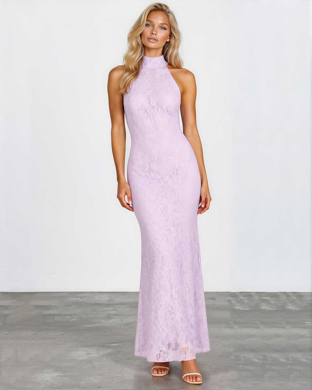 A woman modeling a sleeveless, high-neck, floor-length lavender lace maxi dress against a white studio wall and gray floor.