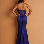 A woman wearing a strapless royal blue maxi dress, shown from the back against a solid tan studio background. The dress features a lace-up open back and a fitted silhouette.