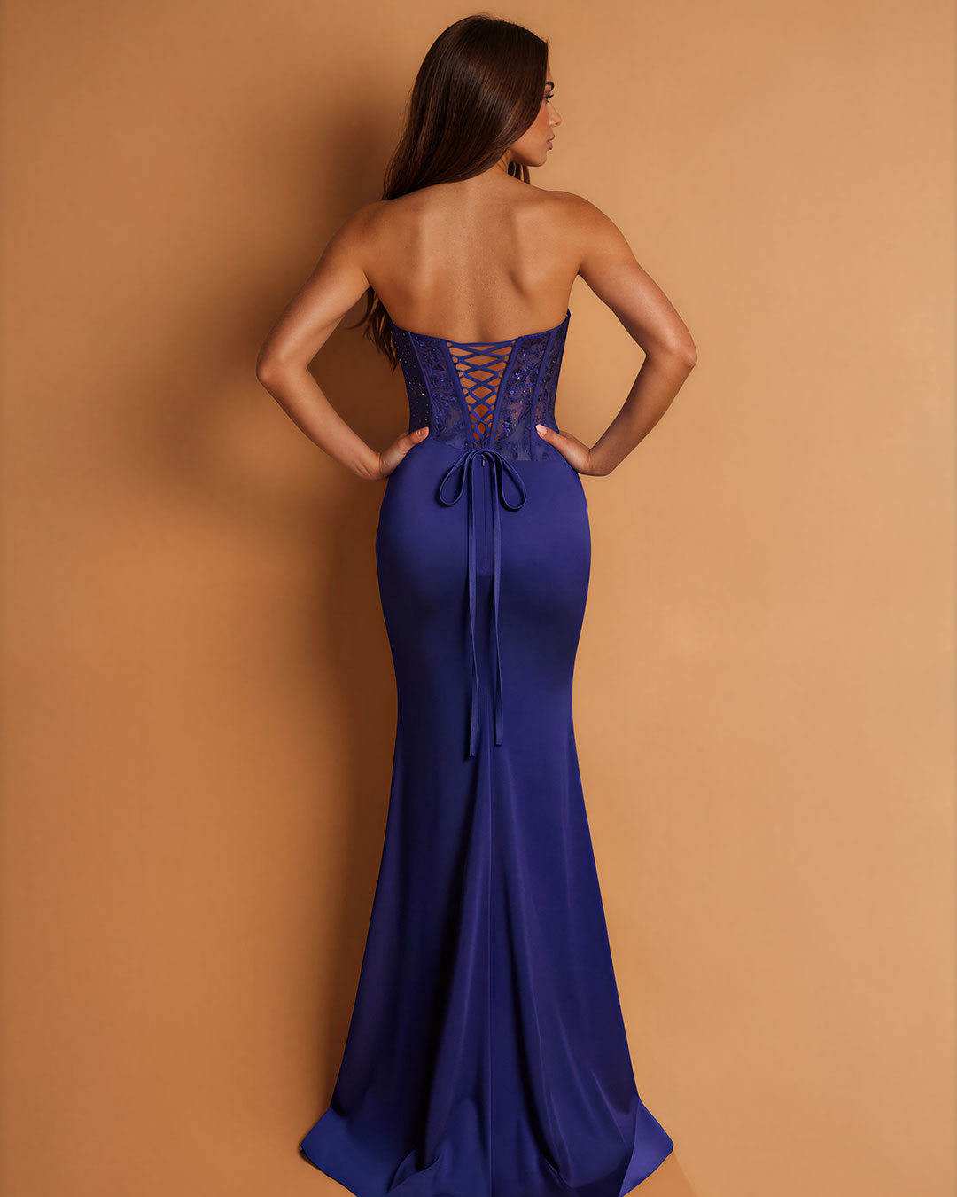 A woman wearing a strapless royal blue maxi dress, shown from the back against a solid tan studio background. The dress features a lace-up open back and a fitted silhouette.