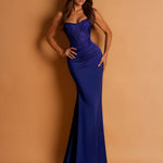 A woman modeling a strapless royal blue evening gown with a heavily embellished corset-style bodice and a fitted mermaid silhouette, posed against a warm tan studio backdrop.