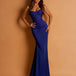 A woman modeling a strapless royal blue evening gown with a heavily embellished corset-style bodice and a fitted mermaid silhouette, posed against a warm tan studio backdrop.