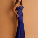 A woman modeling a strapless royal blue evening gown with a fitted corset-style bodice and flared skirt, posed against a warm tan studio backdrop.