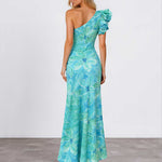 A woman modeling a one-shoulder maxi dress with a vibrant blue and green floral print, shown from the back against a plain studio background.