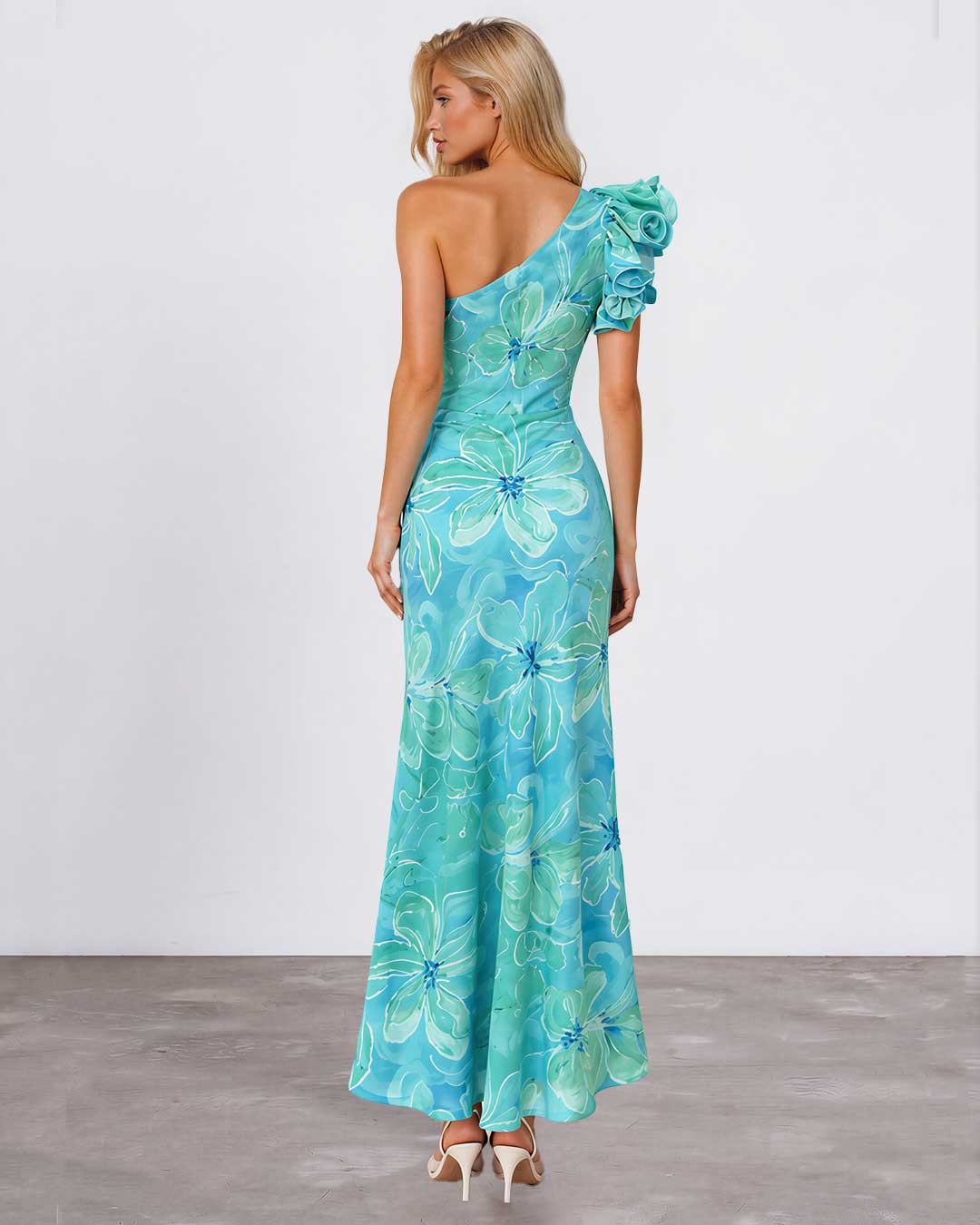 A woman modeling a one-shoulder maxi dress with a vibrant blue and green floral print, shown from the back against a plain studio background.