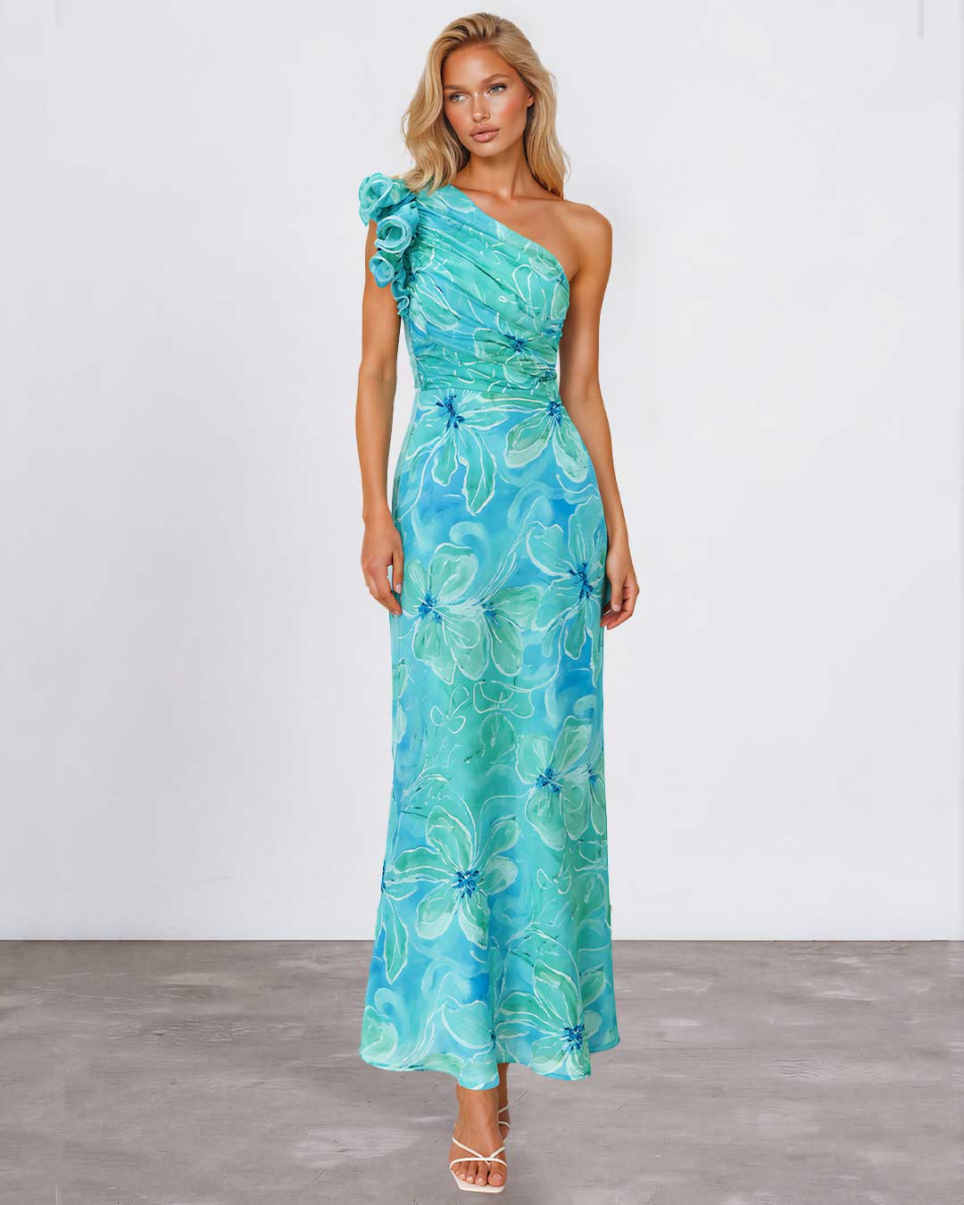A woman modeling a one-shoulder maxi dress with a vibrant turquoise and teal floral print, standing against a neutral studio background.