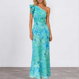 A woman modeling a one-shoulder maxi dress with a vibrant turquoise and teal floral print, standing against a neutral studio background.