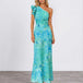 A woman modeling a one-shoulder maxi dress with a vibrant turquoise and teal floral print, standing against a neutral studio background.