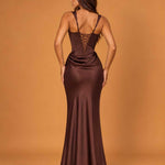 A woman wearing a floor-length satin-like gown shown from the back against a warm tan studio backdrop. The dress is a deep chocolate brown with a lace-up open back and fitted silhouette.