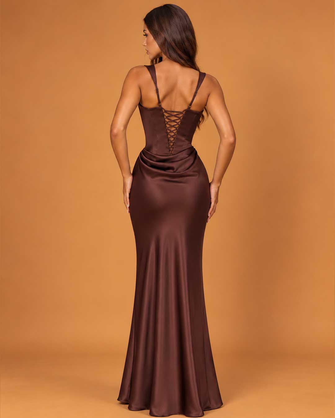 A woman wearing a floor-length satin-like gown shown from the back against a warm tan studio backdrop. The dress is a deep chocolate brown with a lace-up open back and fitted silhouette.