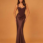 A woman modeling a floor-length, satin-like dark brown gown with a sweetheart neckline, structured bodice, draped waist, and fitted mermaid-style skirt against a warm tan studio backdrop.