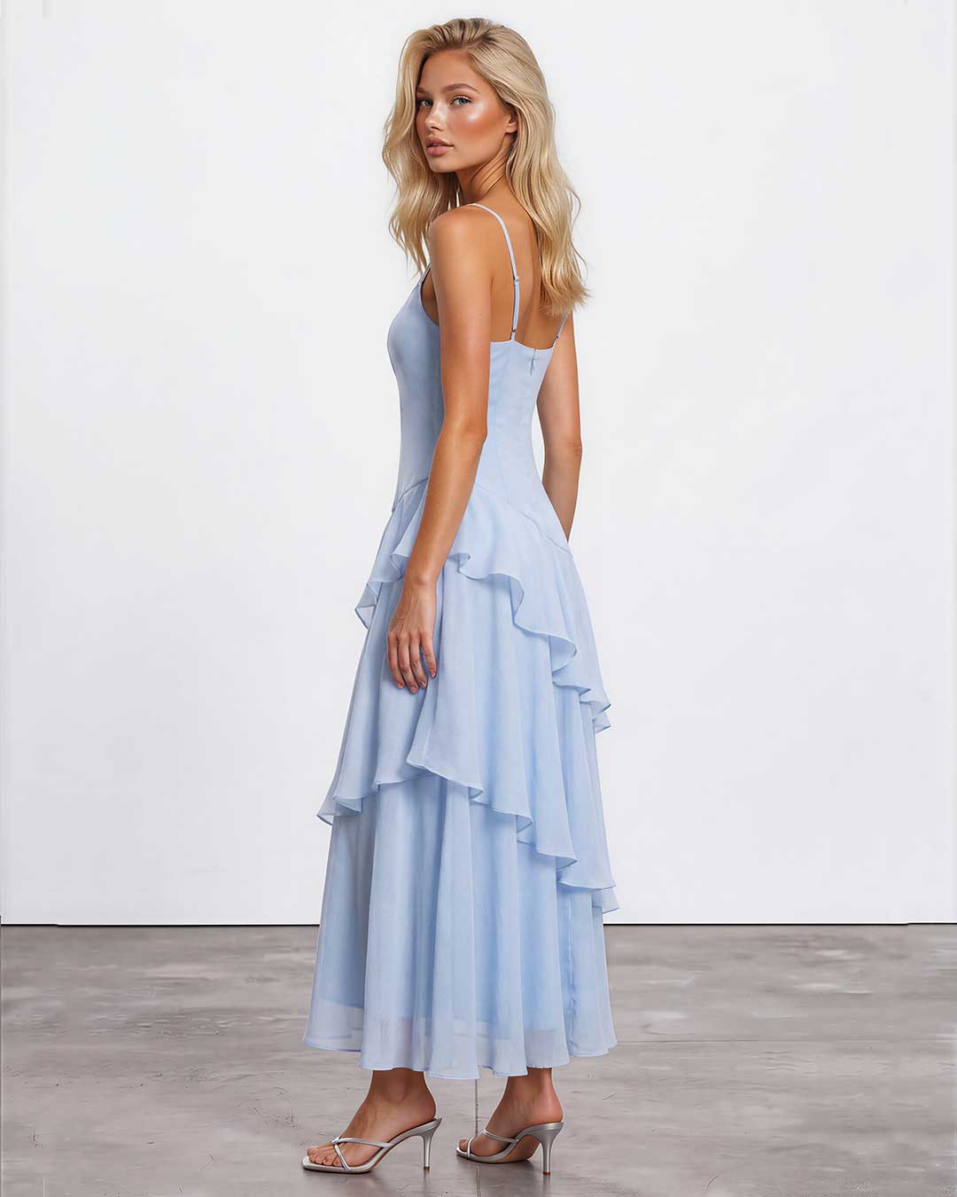 A blonde model wearing a light blue spaghetti-strap maxi dress with a ruffled tiered skirt, shown from a three-quarters back angle against a white studio wall and gray concrete floor.
