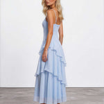 A blonde model wearing a light blue spaghetti-strap maxi dress with a ruffled tiered skirt, shown from a three-quarters back angle against a white studio wall and gray concrete floor.