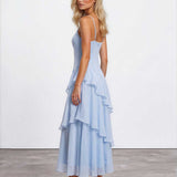 A blonde model wearing a light blue spaghetti-strap maxi dress with a ruffled tiered skirt, shown from a three-quarters back angle against a white studio wall and gray concrete floor.