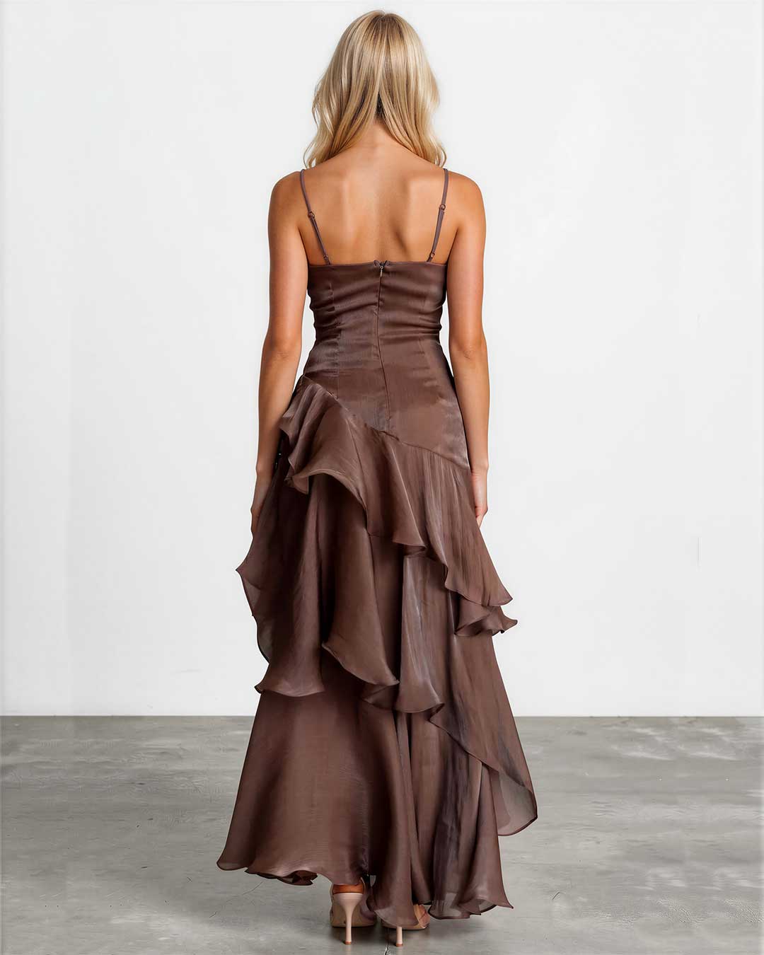 A woman viewed from the back wearing a brown spaghetti-strap maxi dress with a ruched corset-style bodice and a layered ruffled skirt, standing against a plain light gray studio wall and concrete floor.