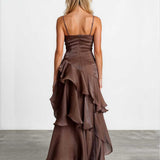 A woman viewed from the back wearing a brown spaghetti-strap maxi dress with a ruched corset-style bodice and a layered ruffled skirt, standing against a plain light gray studio wall and concrete floor.