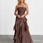 A woman modeling a brown strapless maxi dress with a ruched bodice and layered ruffle skirt, standing against a light gray studio wall.