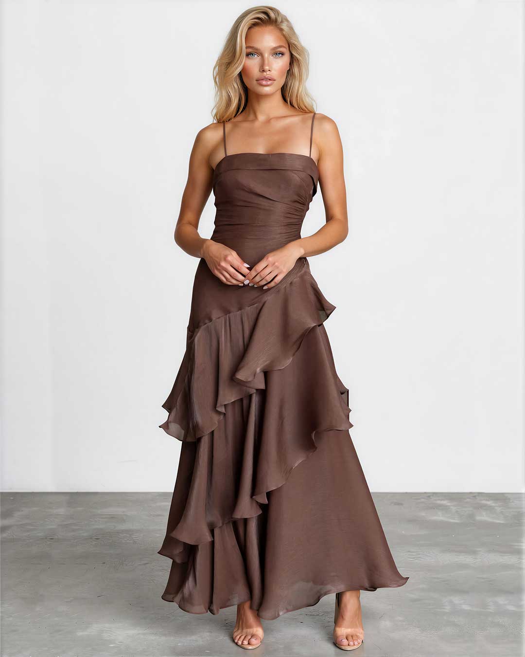 A woman modeling a brown strapless maxi dress with a ruched bodice and layered ruffle skirt, standing against a light gray studio wall.